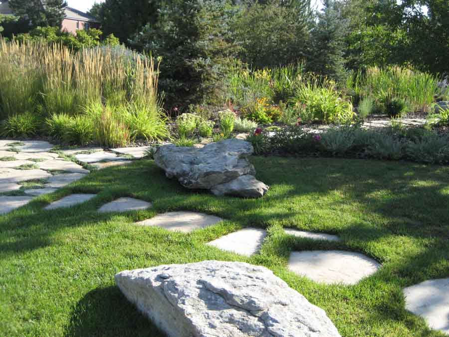 Flagstone path across a lawn past boulders toward ornamental grasses and perennial beds